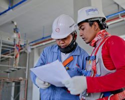 Two engineers in safety helmets reviewing construction plans at a worksite.